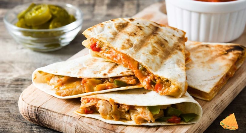 Chicken quesadilla slices garnished with fresh pico de gallo on a wooden tray, served with tomatoes and pickles