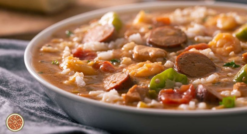 Gumbo highlighting shrimp, sausage, and colorful vegetables, served over white rice in a rich, savory sauce.