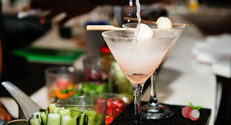 Refreshing lychee martini being poured into a cocktail glass with garnished lychees, set against a vibrant bar background