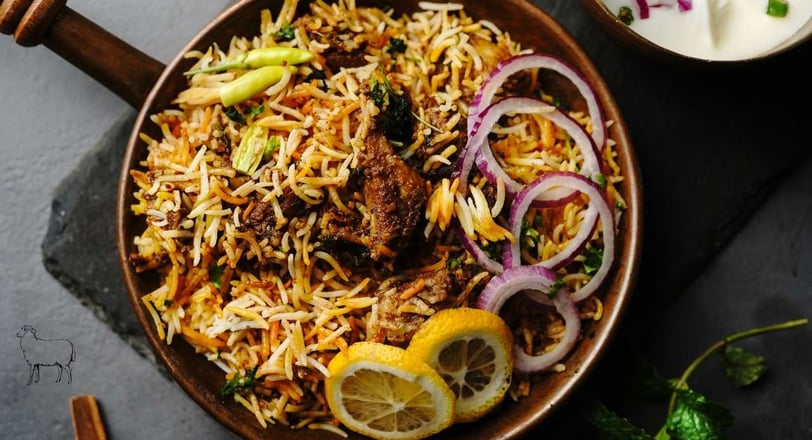 Mutton biryani garnished with onions, lemon, and chilies in a copper pot on a slate background