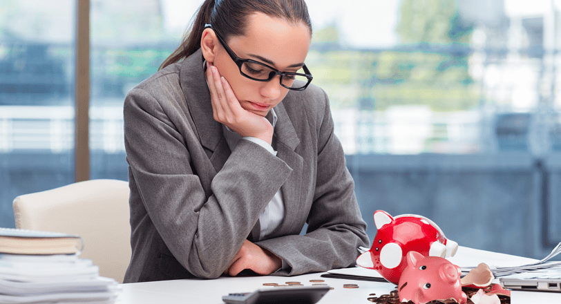 A sad woman sitting at a desk looking at her broken piggy bank and coins