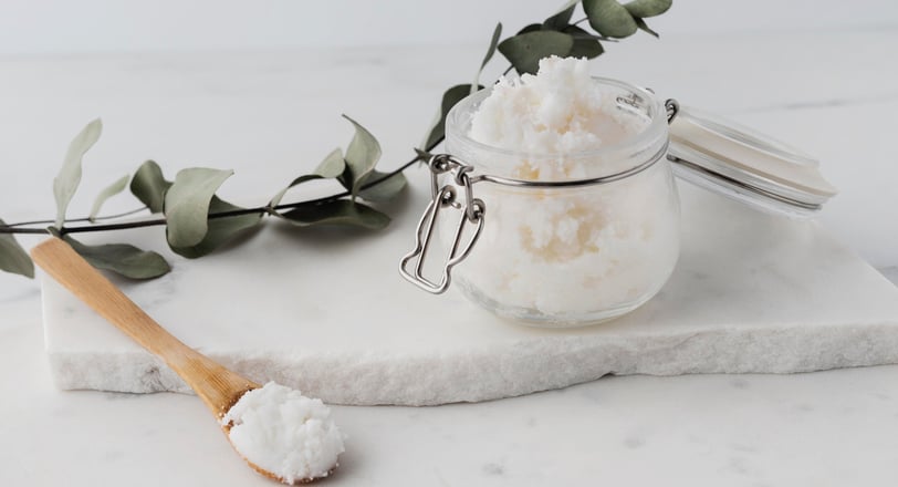 a spoon and a glass jar with a white scrub in it sitting on a marble slab and a eucalyptus leaf