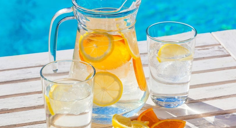 a jug and two glasses with water, ice and lemon and orange slices with a swimming pool in the backgr