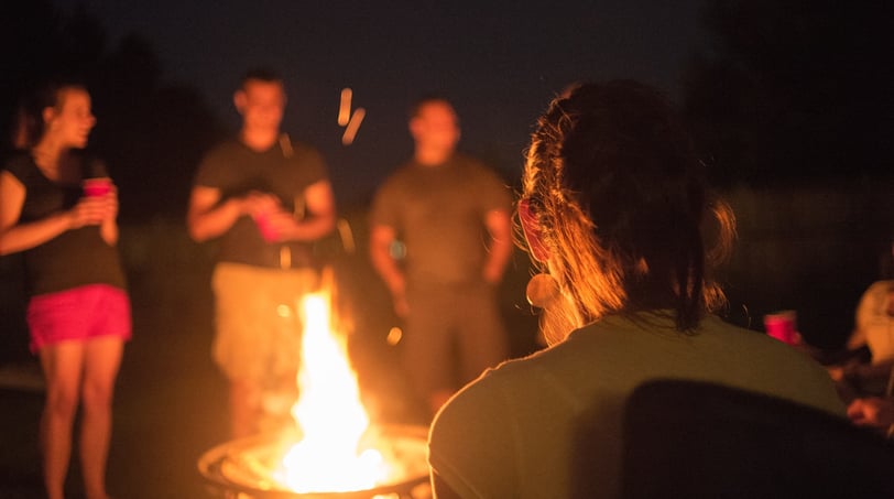 A nighttime gathering around a glowing campfire, with silhouettes of people standing in a circle. The warm firelight illumina