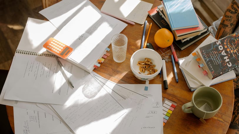 A cluttered round table, containing paper, crockery, food, books and stationary.