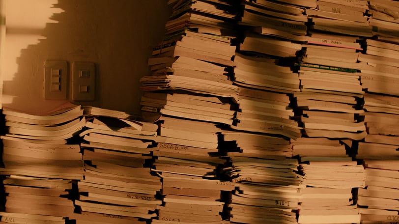 Multiple piles of books, stacked agains a wall. A pair of lightswitches is visible beside the piles.