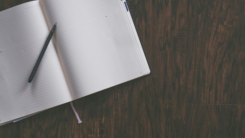 An open lined notebook on a wooden table, with a pen laid across it.