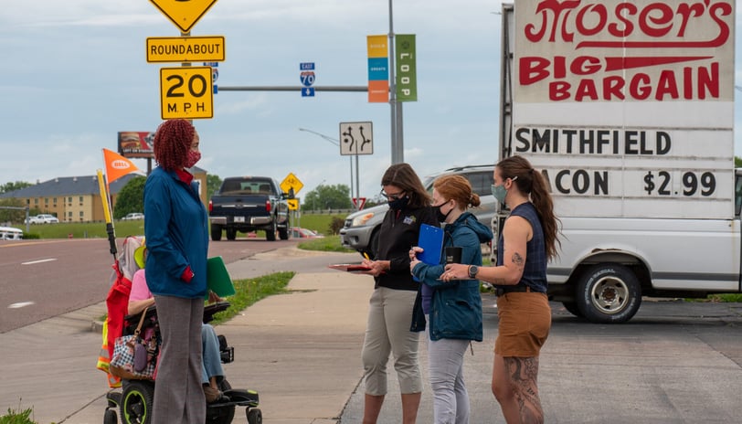 Felecia on a sidewalk next to a busy road speaking to a group of people with clipboards