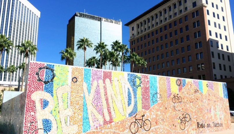 Big city picture with palm trees, tall buildings, and a mural spelling out "Be Kind" with bikes.