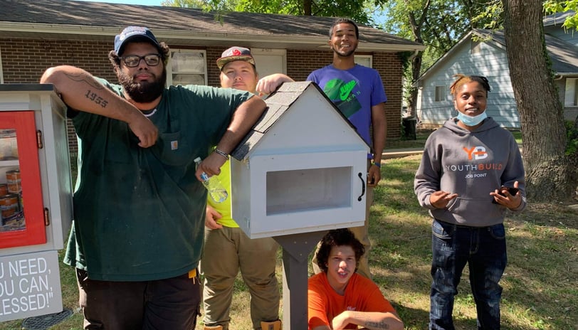 A group of kids and adults with Matthia, posing with a little library in front of a house