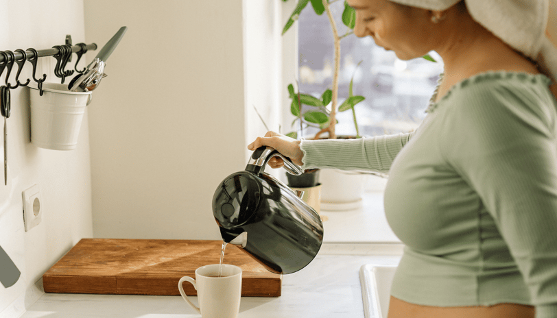 Woman in towel and green top pouring tea in sunlit kitchen