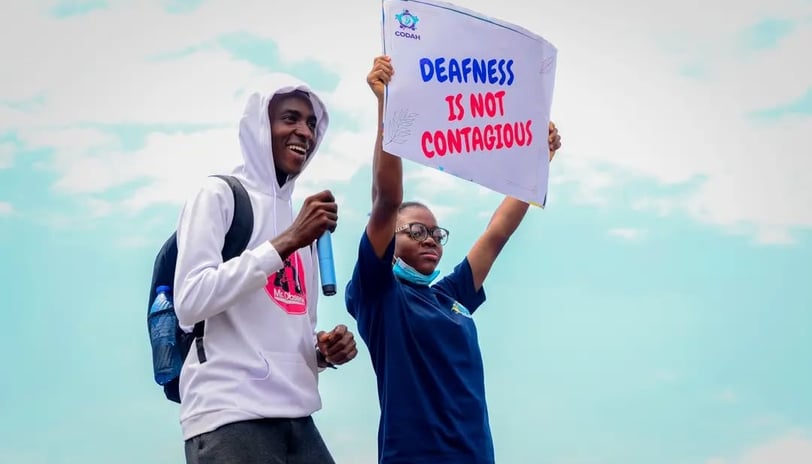 A man in a white hoodie, standing next to a woman who is holding up a banner that says "Deafness is not contagious"