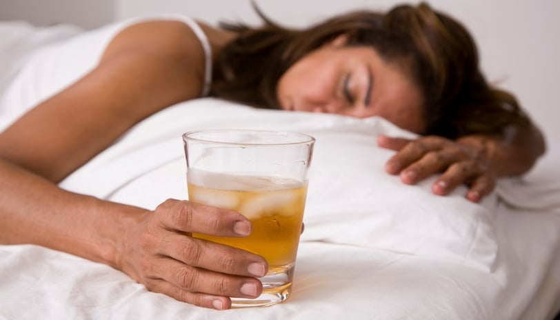 A woman looking unwell lies in bed while holding a glass containing an alcoholic drink, depicting the negative consequences 