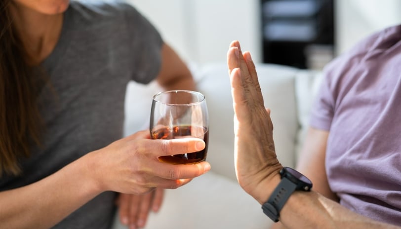A person holds up their hand in a clear "stop" gesture, refusing a glass of dark liquor being offered to them. 