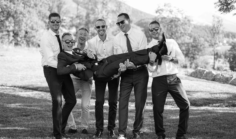 Photo en noir et blanc d'un marié porté par ses amis lors d'une séance de photos de groupe.