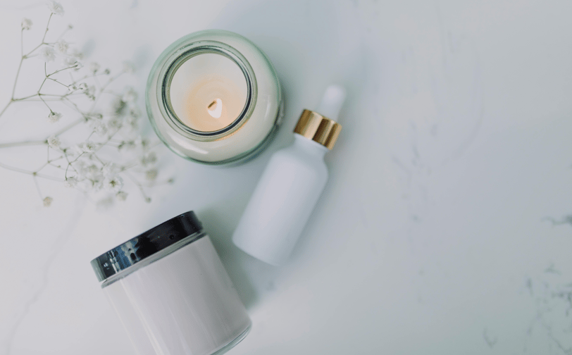 Image showing from above a white candle, a glass container of a moisturizer and a serum, all placed on a white marble surface