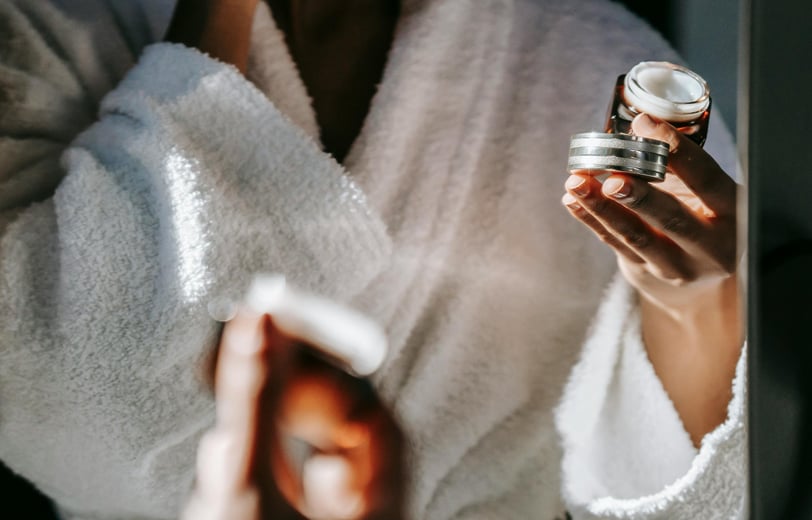 A woman looking at a mirror, wearing an eye cream with one hand and holding the cream container on the other hand.