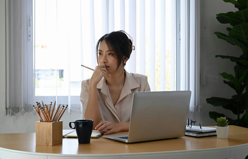 A thoughtful businesswoman in her office, sitting at her desk with an open laptop computer.
