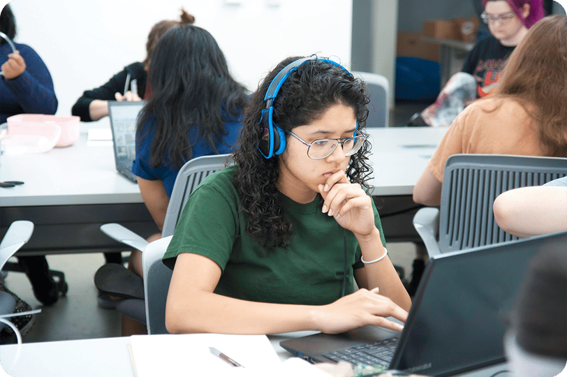 Mulher jovem de óculos e fones de ouvidos, utilizando notebook sobre a mesa, em uma sala de aula