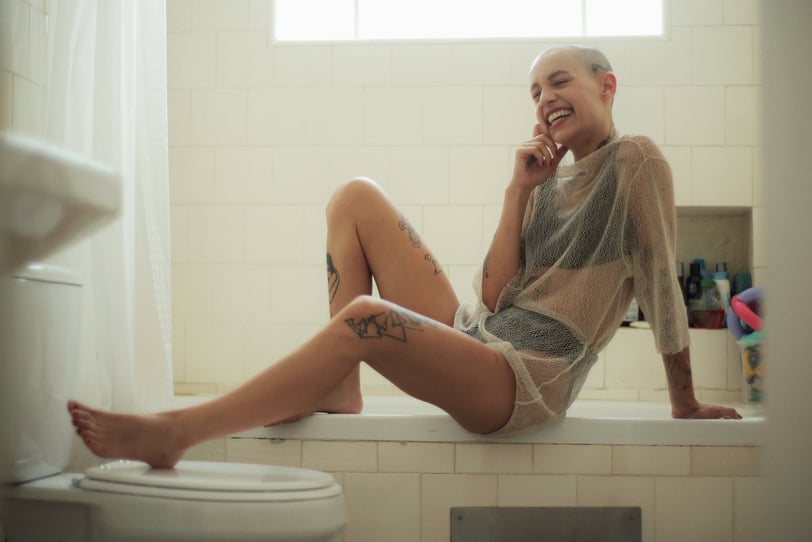 A woman sitting in the bathroom, on the lip on her bathtub, smiling and touching her face