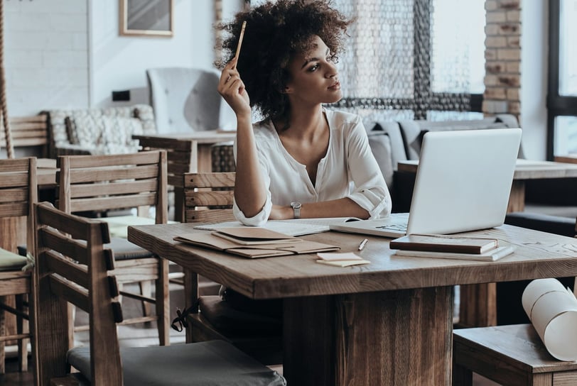A thoughtful young woman sitting at a table in front of an open laptop.