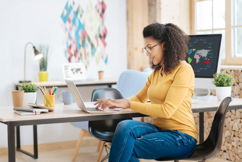 a woman wearing a mustard sweater and jeans, sitting at a desk working on a laptop computer