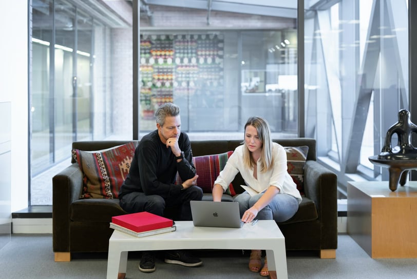 A man and woman sitting on a sofa in an office, looking at a Macbook.