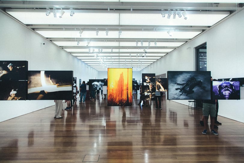Pessoa admirando uma pintura em uma galeria de arte com iluminação suave e obras em exibição ao fund