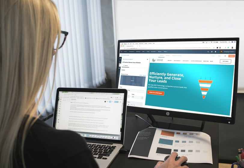 a woman sitting at a desk with a laptop and a monitor showing an email marketing platform