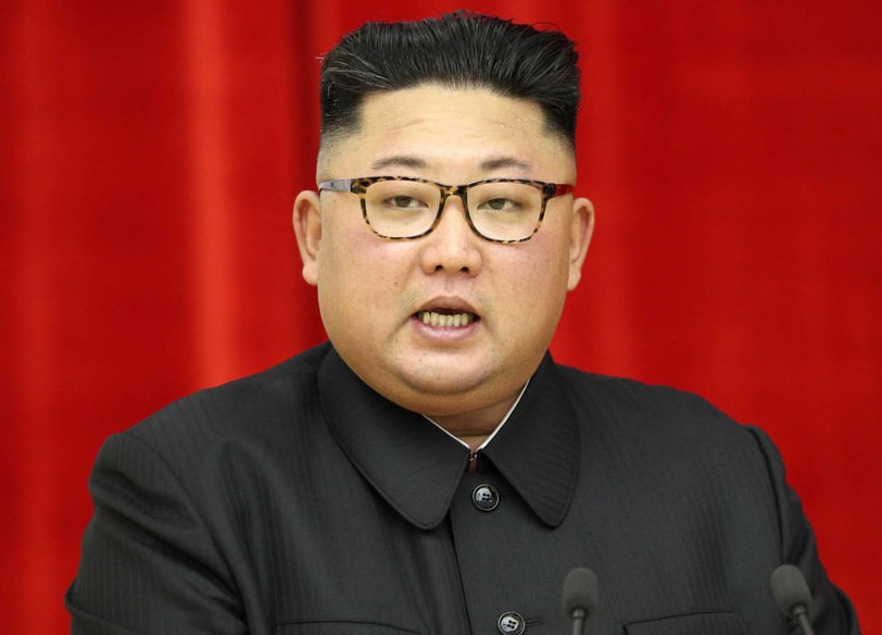 Kim Jong-un in a suit and glasses is standing Infront of a microphone