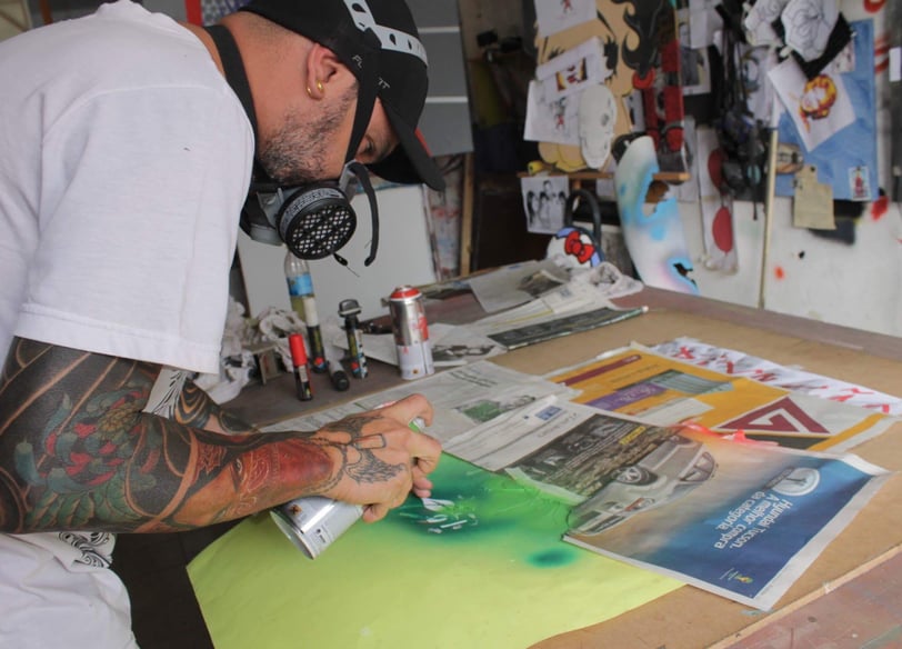 Detalhe das mãos do artista enquanto ajusta spray e tintas sobre a mesa de trabalho.