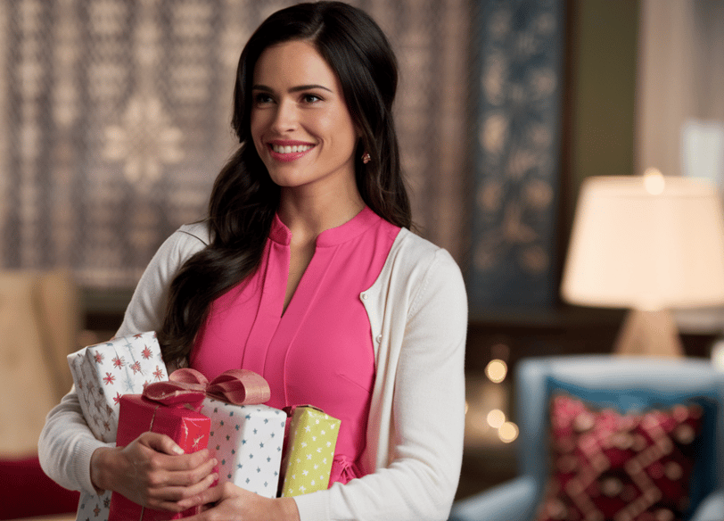 Une femme sublime, souriante, habillée d'une robe rose. Elle porte plein de cadeaux Noel 2024