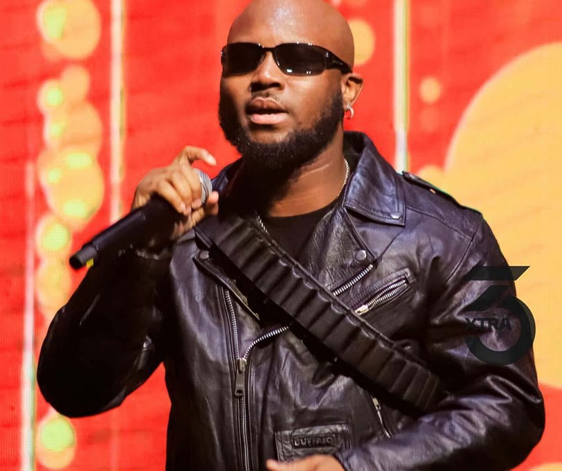 King Promise in a black jacket in sunglasses with a microphone.