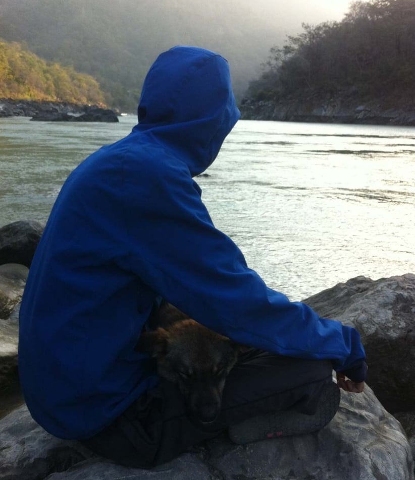 a man in a blue jacket sitting on a rock by the side o river ganga with dog sleeping in his lap