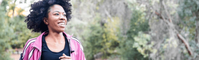 Une jeune femme noire souriante aux cheveux naturels et une veste de sport rose qui fait son jogging