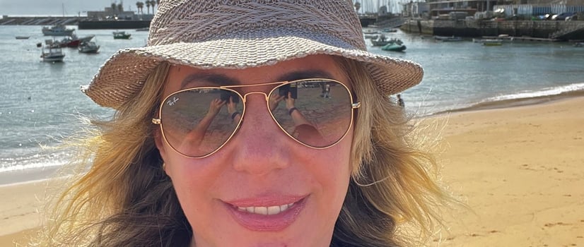 autora com chapéu e óculos de sol em praia com barcos ao fundo