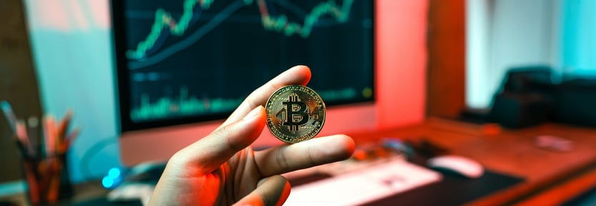 A person holds a bitcoin in front of a computer screen displaying cryptocurrency data and charts.