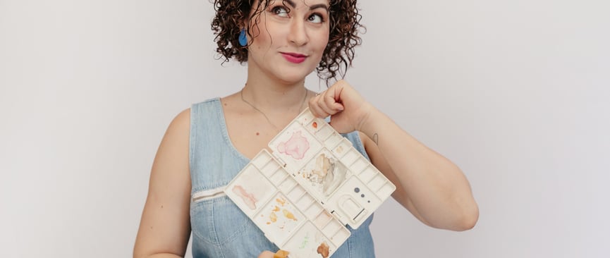 Uma mulher de vestido azul e cabelo cacheado segurando uma paleta de aquarela