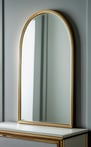 A thin gold-framed arch mirror leans elegantly against a grey wall atop a wooden dresser.