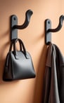 Two matte black wall hooks holding a black handbag and grey coat against a peach wall.