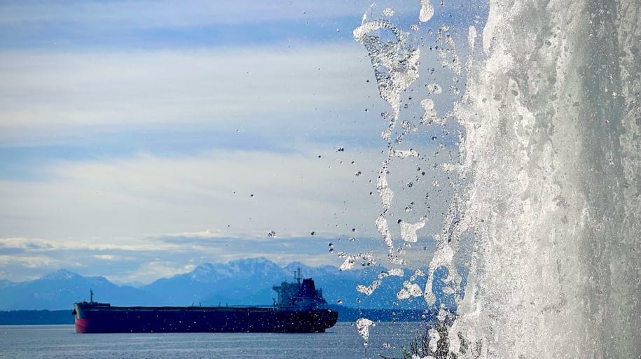 A fountain in front of a cargo ship in the Puget Sound with mountains in the background