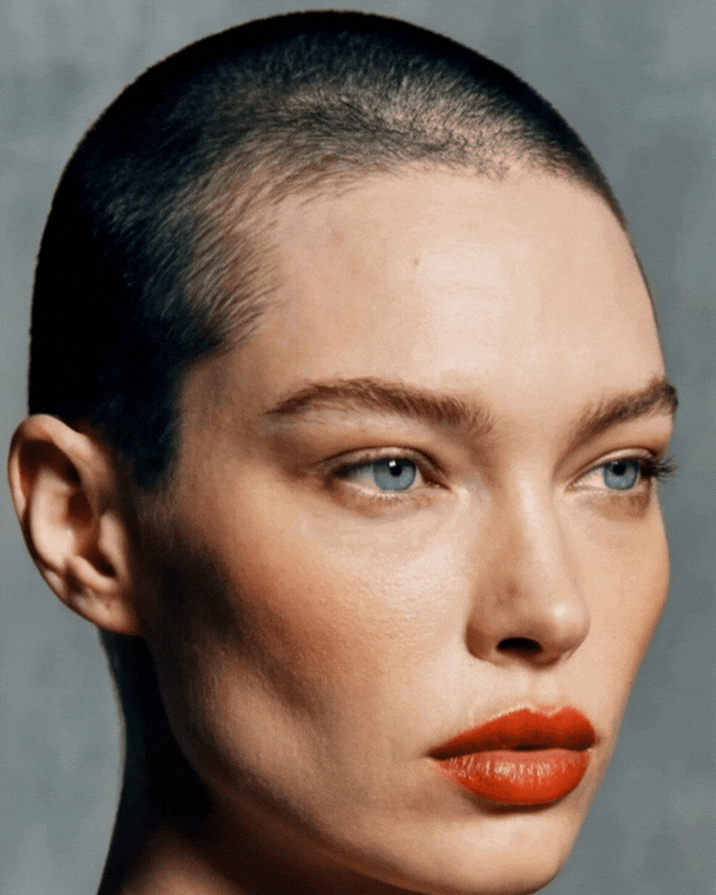 A woman with a buzz cut hairstyle, bold red lipstick, and blue eyes posing for a beauty portrait.