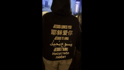 Person in black hoodie at night, city lights behind, back reads Jesus loves you in many languages.